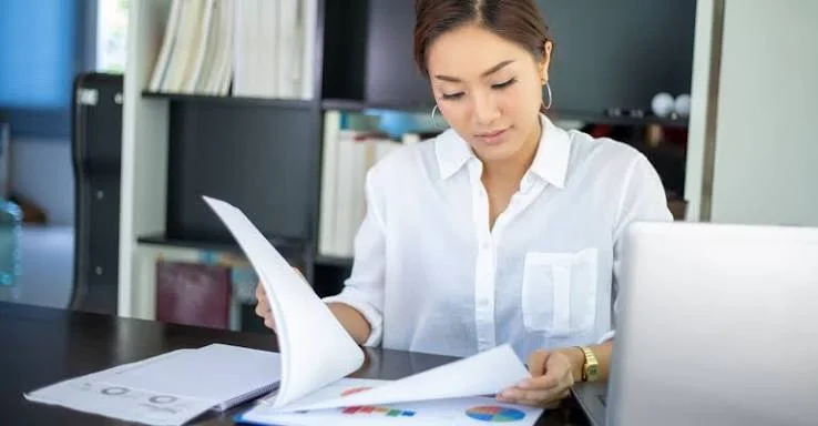 A contractor at her desk reviewing printed statements beside a laptop.